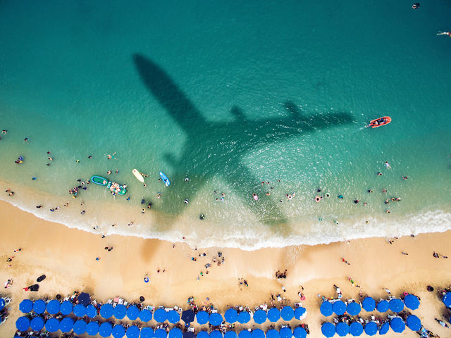 Aerial view of a blue-green beach with swimmers and the shadow of an airplane.